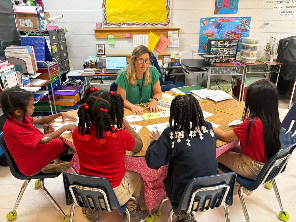 A teacher sits at a table with four students, working with phonics flashcards