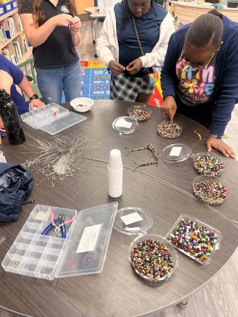 Students stand around a table making bracelets with beads and fishing line