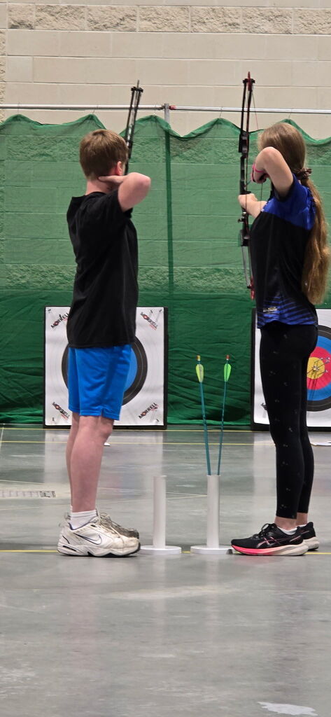 Two individuals are standing with archery bows, aiming at a target. The target has multiple colored rings.