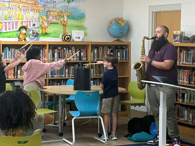 A man plays a saxophone while students play a plastic drum with drumsticks