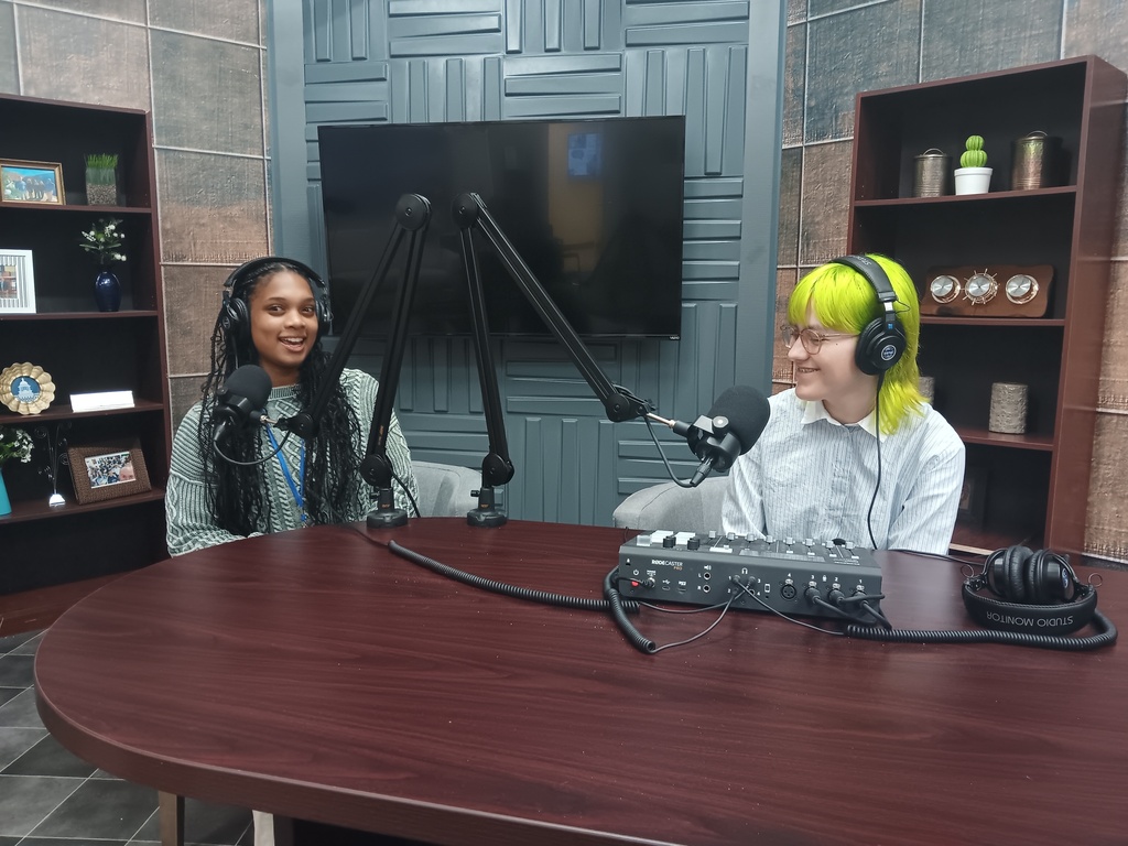 Two high school students sitting at a table behind podcasting microphones
