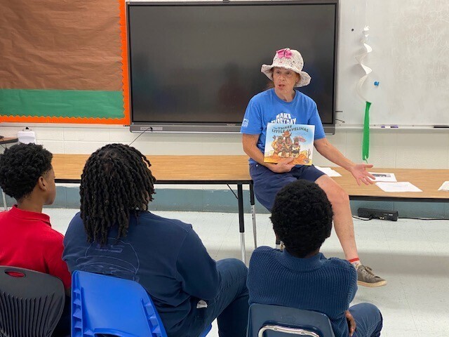 A woman holds a book while speaking to a class of elementary students