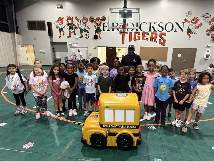 A large group of elementary students stand in a gymnasium with a remote control school bus