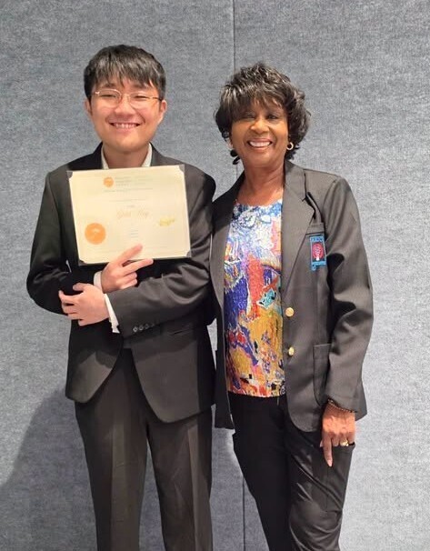 Alex Choi holds a certificate while standing next to a woman in a black blazer