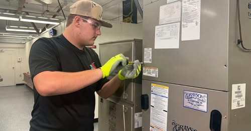 A high school boy in a cap and safety goggles works on an air conditioning unit