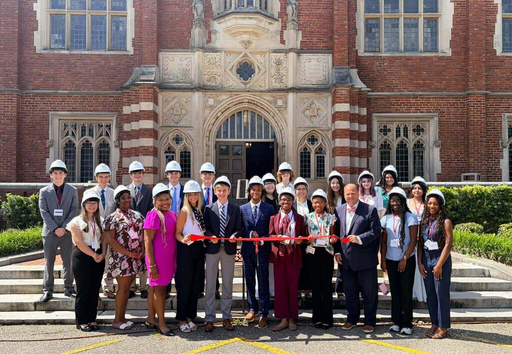 A group of high school students in hard hats, holding a red ribbon
