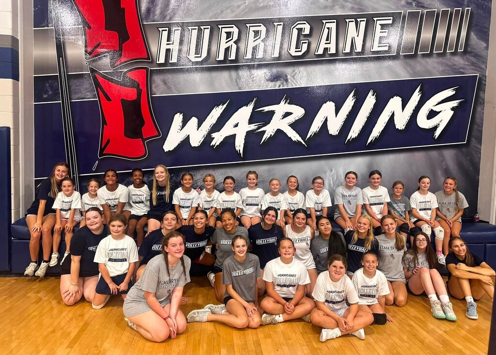 A large group of high school and elementary volleyball players, sitting underneath a mural that reads "Hurricane Warning"