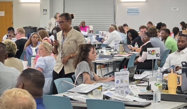 A woman with a microphone walks between tables, where teachers sit with laptops and notebooks