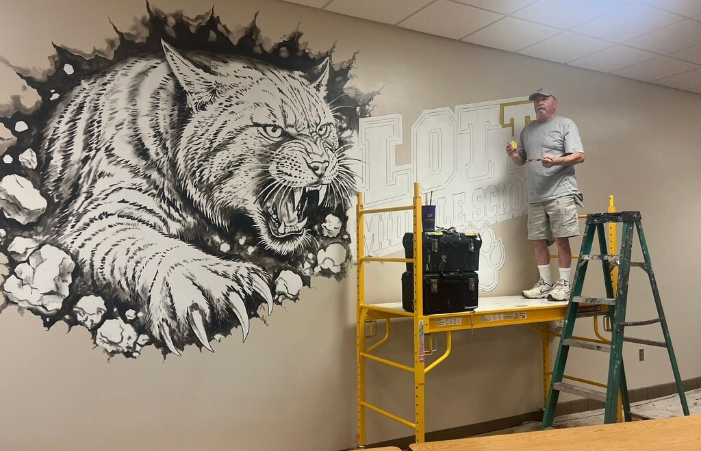 A man on a scaffolding holds a paintbrush next to a mural of a wildcat