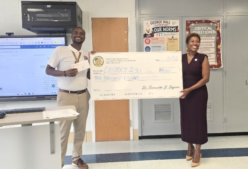 Two people hold a large check in a classroom with posters on the wall and a computer monitor behind them.