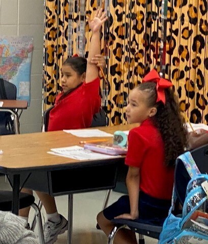 Two students seated at a table, one with her hand raised