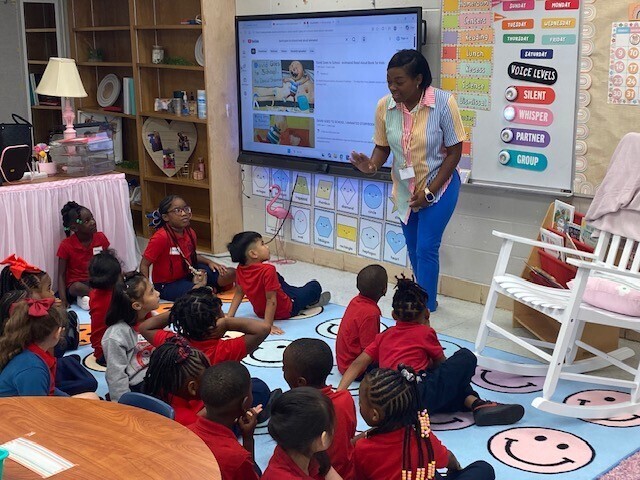 A teacher speaks to students who are seated on a rug in a classroom