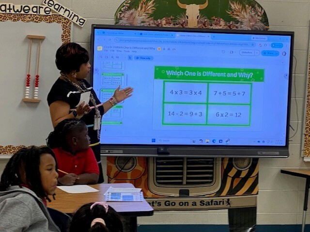A teacher points to multiplication problems on a smartboard