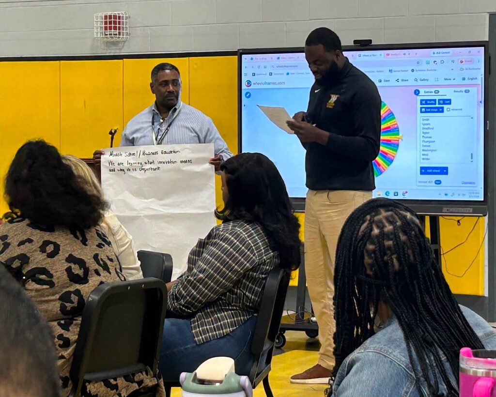 Two men standing in front of a room of teachers, holding a poster with a goal written on it