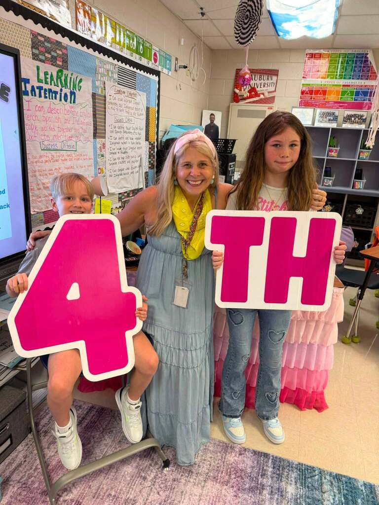A teacher hugs two students while they hold cut outs of the number 4 and the letters TH