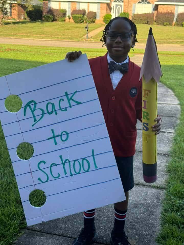 A boy in a red sweater holding an oversized pencil and a sign that says "Back to School"