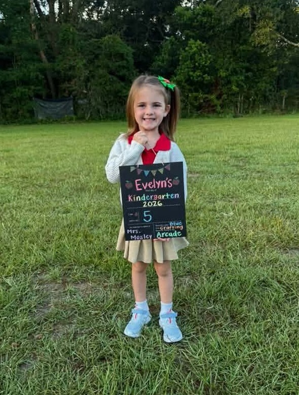 A girl standing outside and holding a sign that reads "Kindergarten 2026"