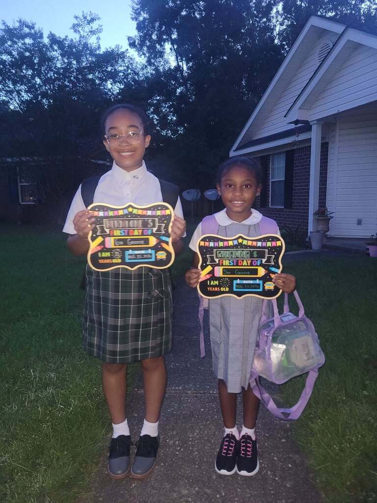 Two girls standing outside a house holding signs and a backpack
