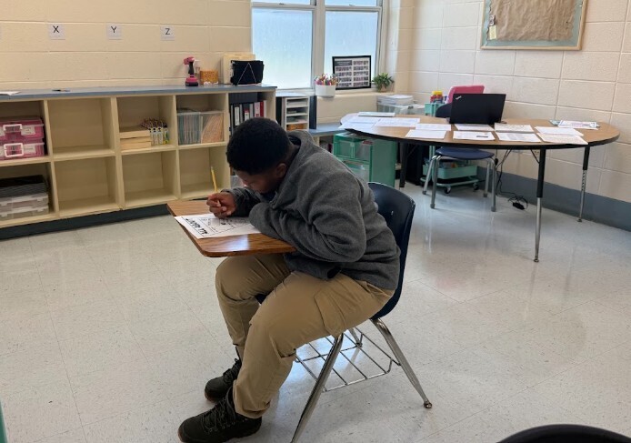 A student in a classroom, sitting on a chair, working on a piece of paper at a desk.