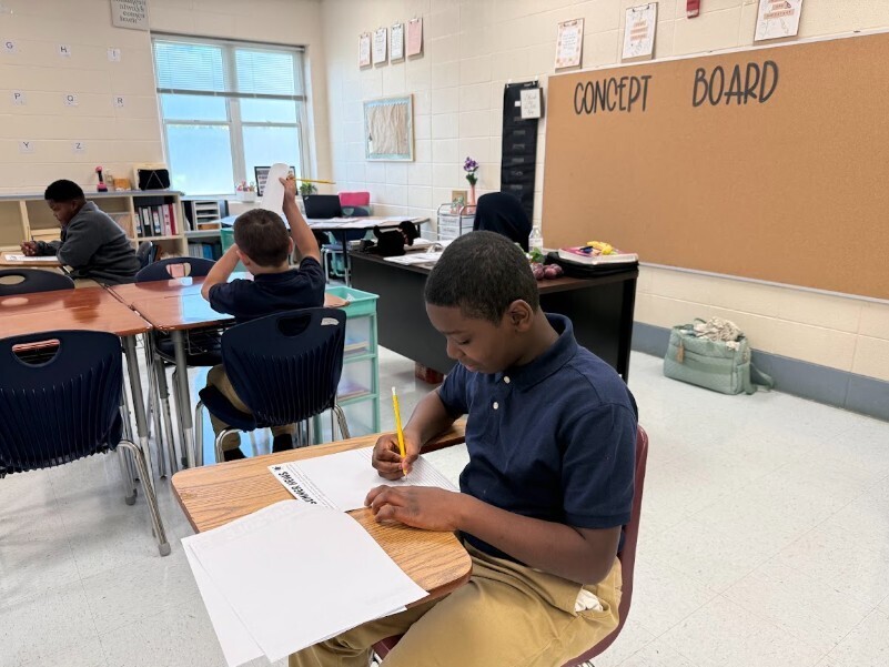 Four students in a classroom setting, seated at desks. One student actively writing, others appear engaged in work.