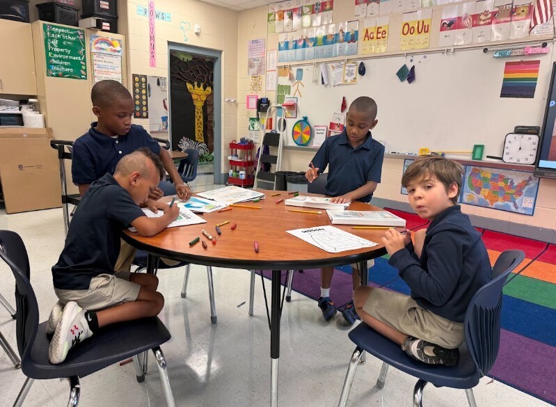 Four children are seated at a round table in a classroom, each with papers and markers. The table is surrounded by chairs, and posters are on the walls.