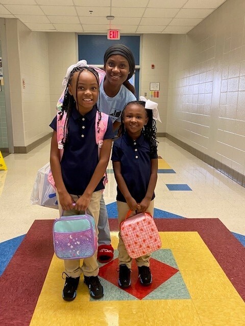 A mother stands with her two daughters, who are holding lunch boxes