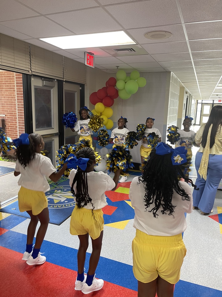 Four girls wearing white shirts, yellow shorts, and blue pom-poms perform in a hallway. A colorful balloon arch is in the background.