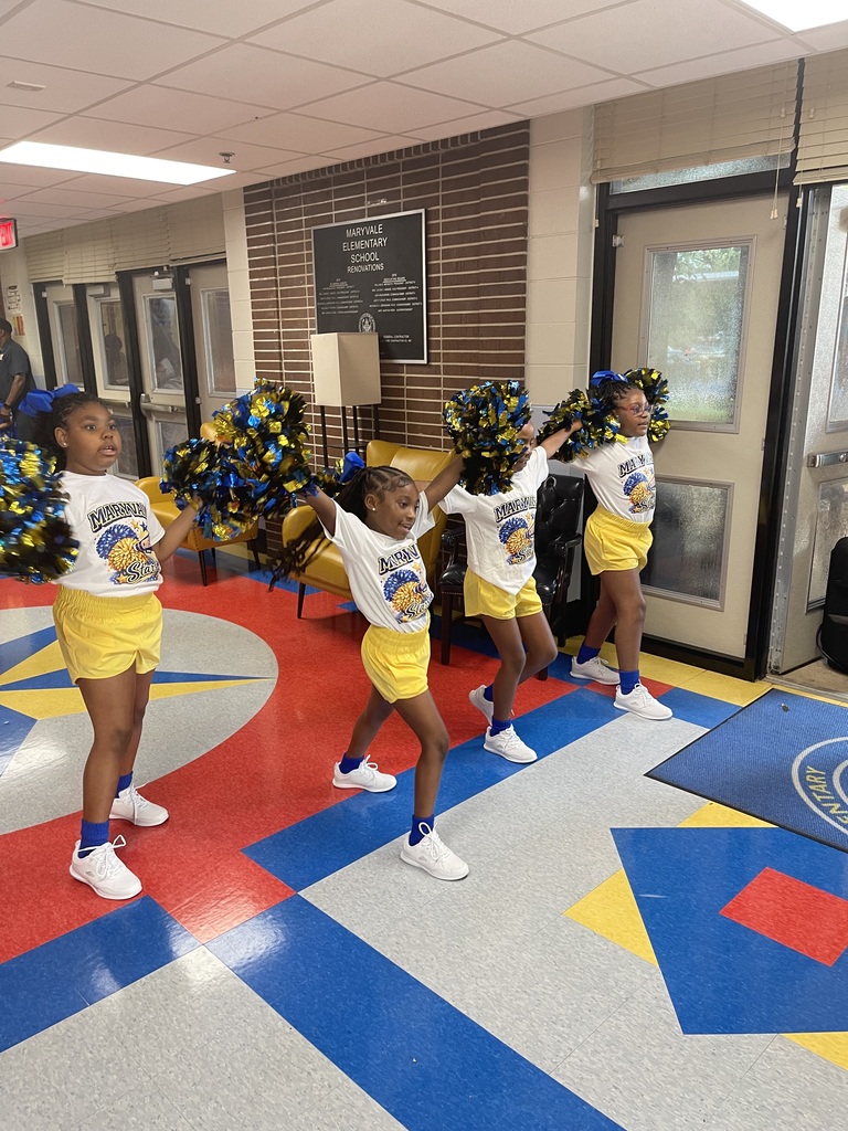 Four young girls in cheerleading uniforms practice a routine in a room with colorful tiles.