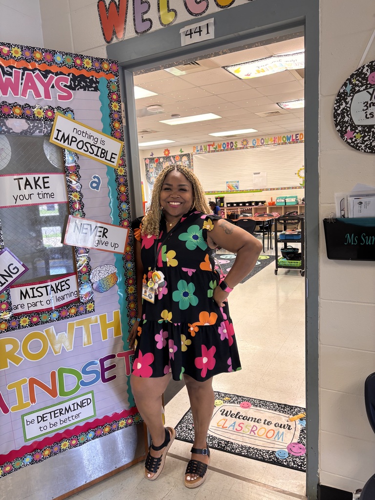 A woman in a floral dress stands next to a bulletin board with inspiring quotes. The room has a white floor and an open door.