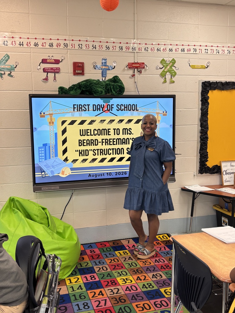 Person stands in front of a classroom screen with "First Day of School" and a welcome message. Colorful rug and chairs are on the floor.