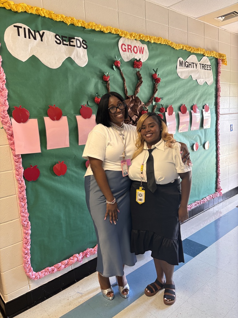 Two women stand in front of a green bulletin board with a tree and red apples.