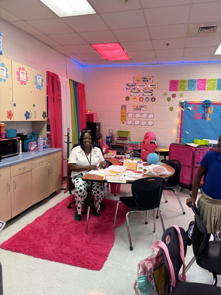 Woman seated at a round table with books and materials, surrounded by chairs. Pink rug, colorful bulletin board, cabinets, and a microwave.