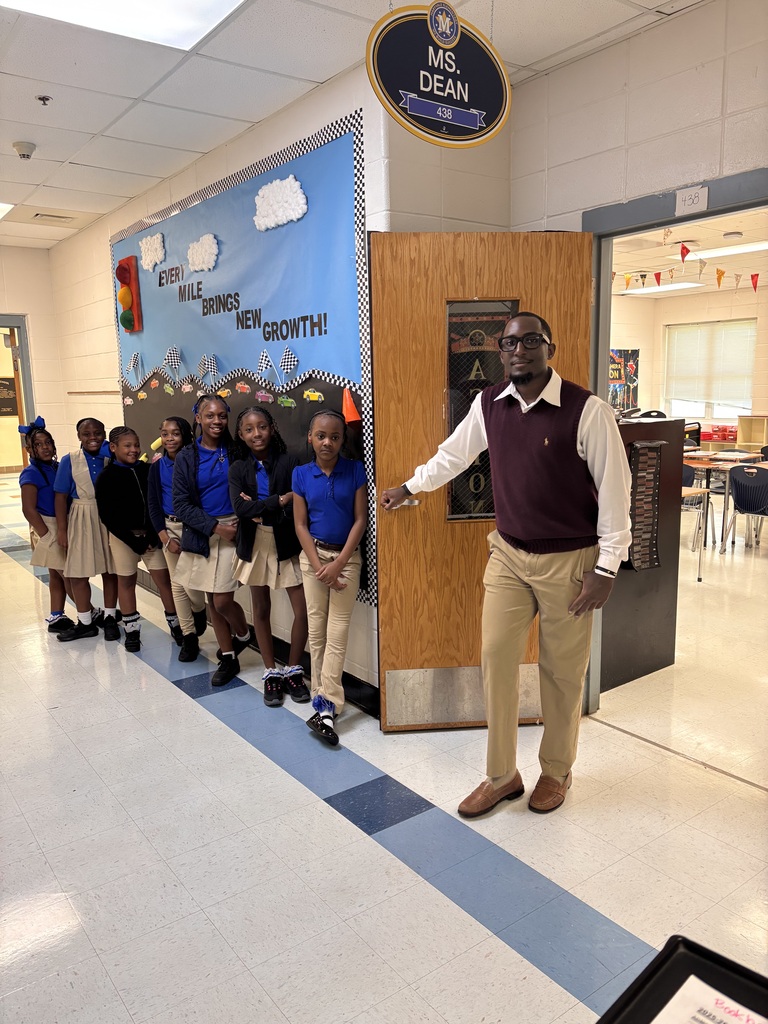 Man in a vest and pants standing next to a wall with young students in blue uniforms.