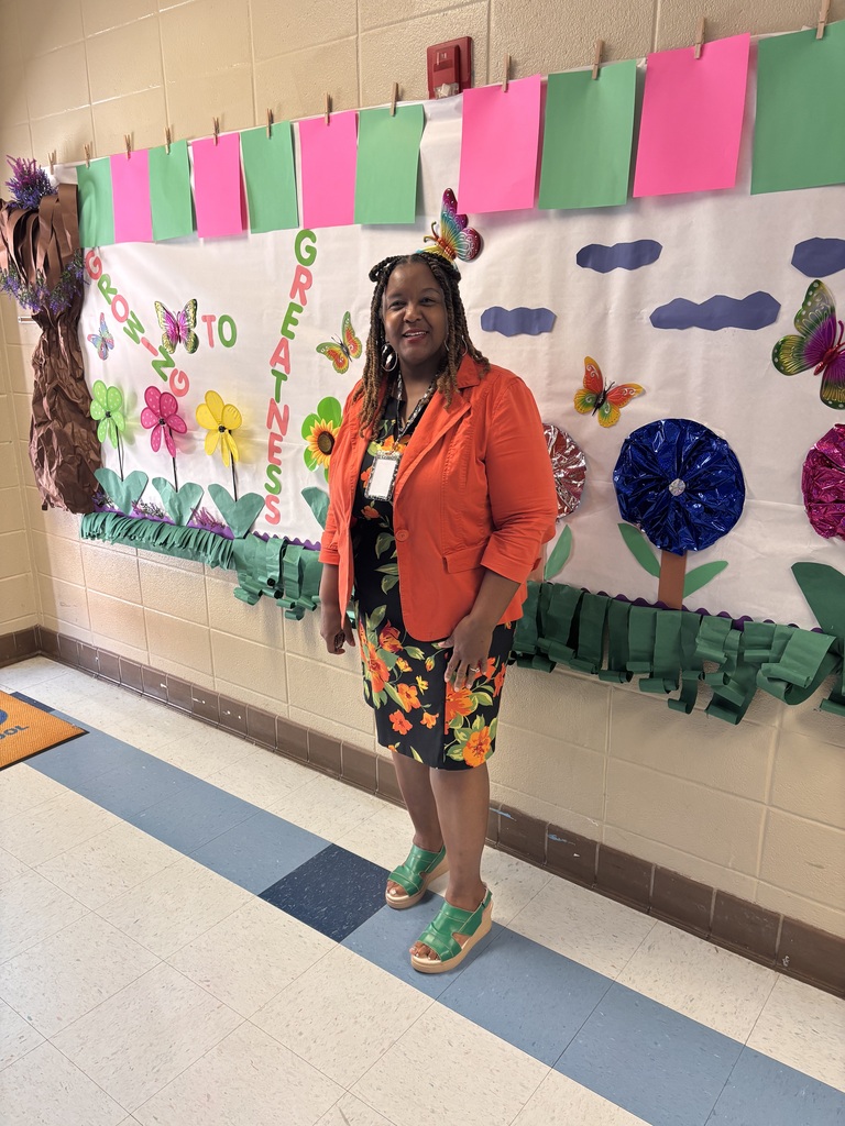 Woman in orange jacket and floral skirt stands in front of a decorated bulletin board in a hallway.