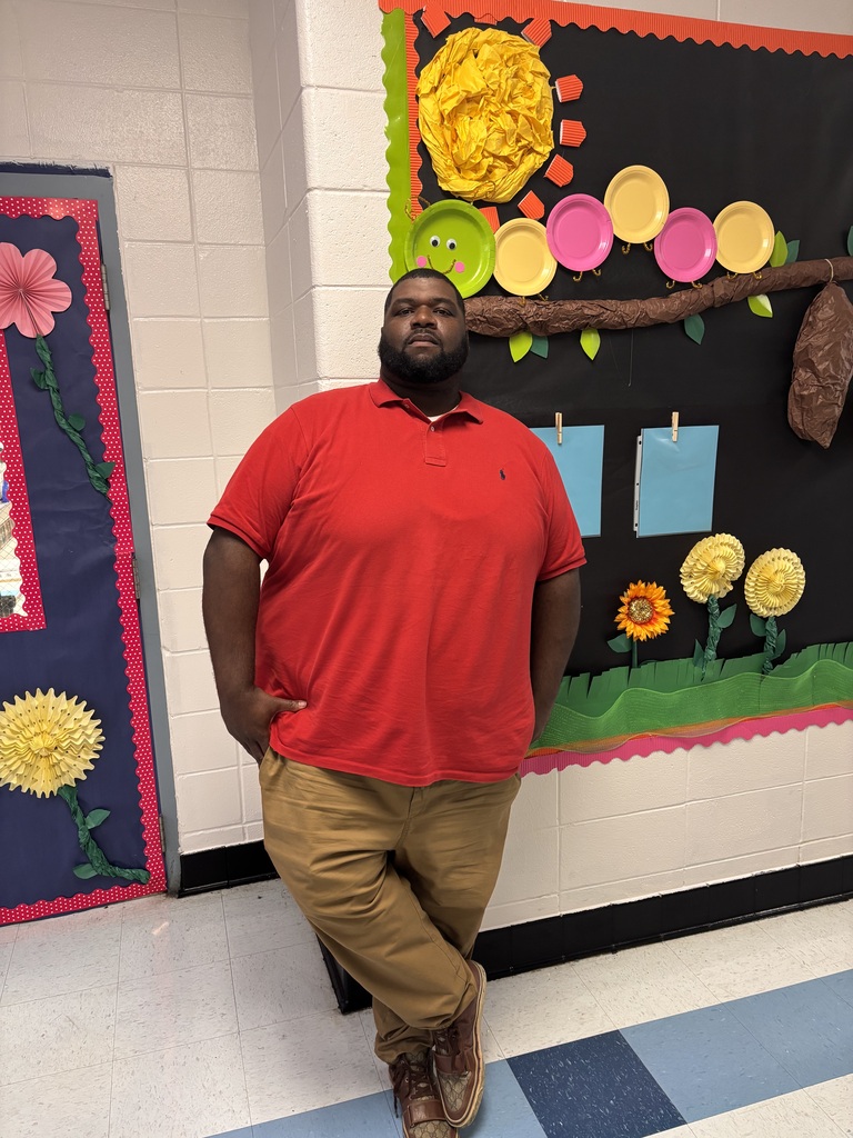 Person in red shirt and brown pants stands in front of a bulletin board with flowers.