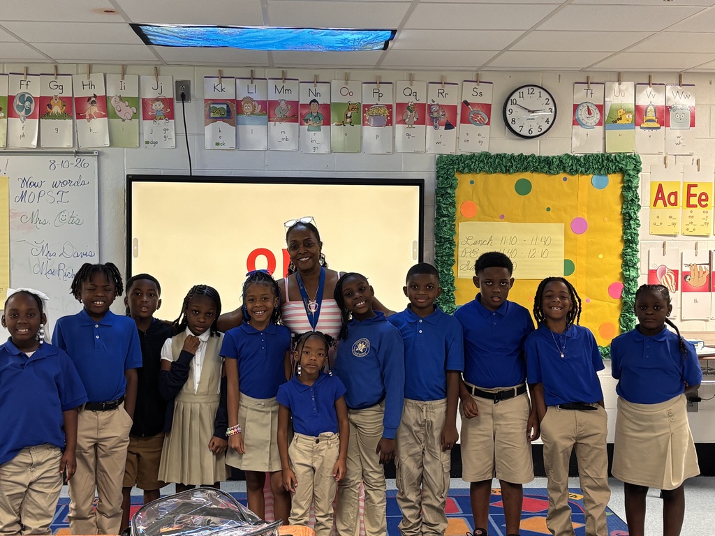 Group of children and a woman in blue uniforms, standing together in front of a screen.