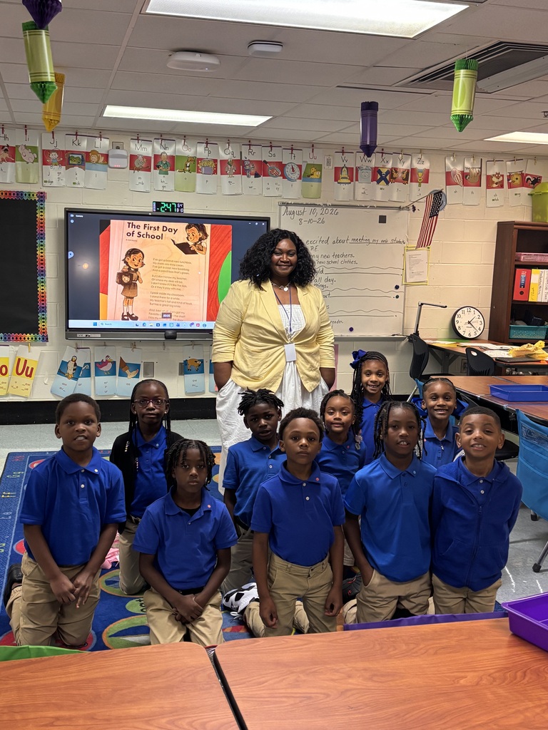 A teacher stands with a group of children wearing blue shirts and khaki pants in a classroom.