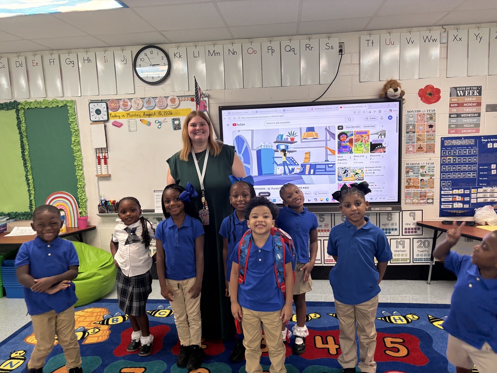 A woman stands in front of a screen showing a presentation. Six children in blue uniforms stand behind her, while others stand to the side.