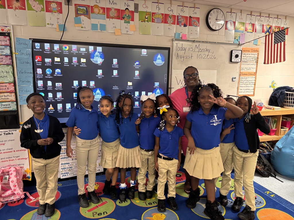 A classroom with a teacher and students, six young girls in matching blue shirts and khaki pants.