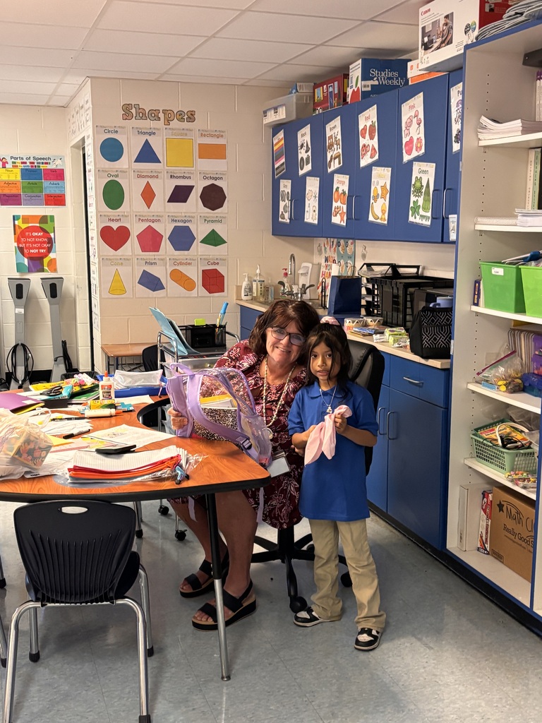 Woman and child standing in a classroom. Woman holds a gift bag. Table with craft supplies.