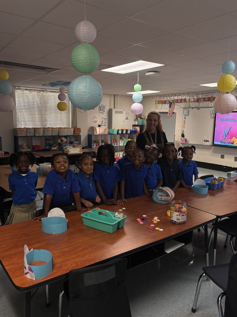 A classroom with children in blue shirts and a teacher standing behind a table with Easter crafts.
