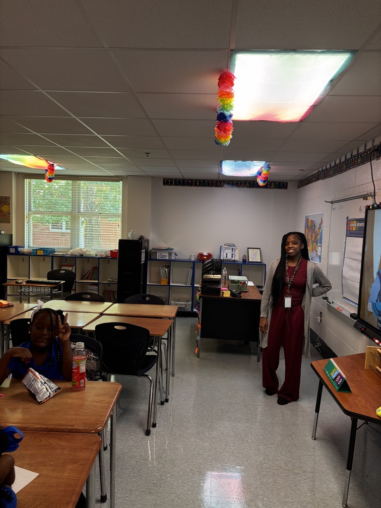 An empty classroom with white walls, tables, and chairs. A person stands in front of a whiteboard. Colorful balloons hang from the ceiling.