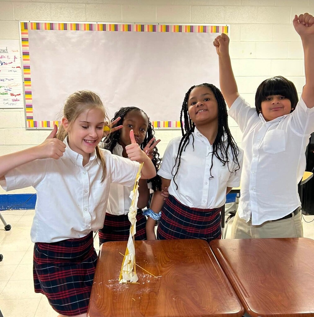 Four students smiling and giving thumbs up beside  a tower made of spaghetti noodles and marshmallows
