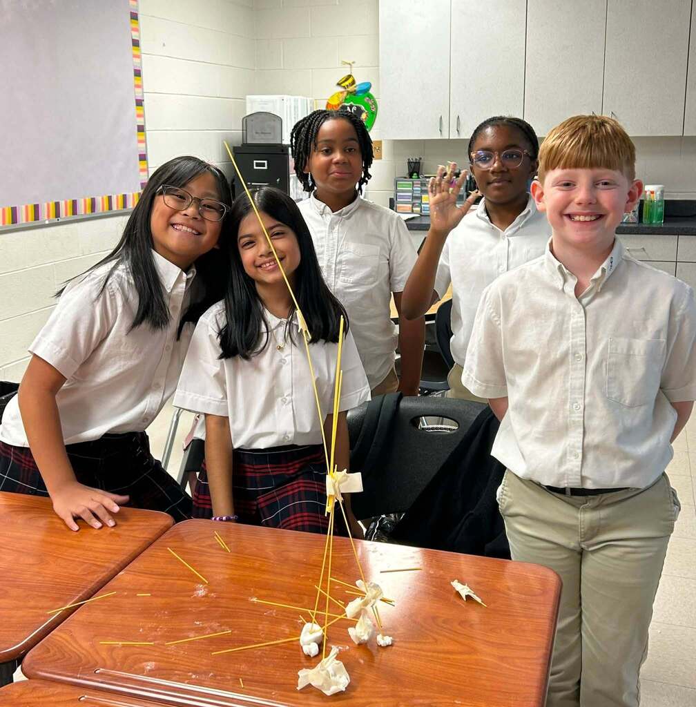Five students smiling beside a tower made of spaghetti noodles and marshmallows