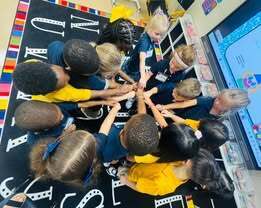 A group of children in blue and yellow shirts forms a circle, holding hands, and looking at a large screen.