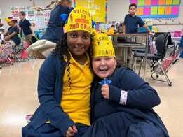 Two children in a classroom setting, wearing yellow hats. One has a blue coat and the other a blue dress.