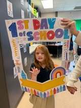 Young girl stands in a school hallway, holding a large cardboard sign that says "1st Day of School."
