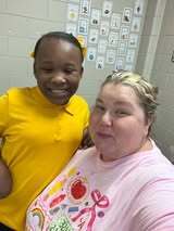 Two women smiling in a room with tiles and cards on the wall. One wears a yellow shirt, the other a pink one with colorful designs.