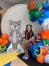 A young girl poses in front of a large backdrop with a cartoon fox and a balloon arch.