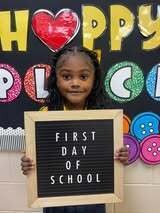 A smiling child holds a sign with the words "FIRST DAY OF SCHOOL". Behind them is a colorful bulletin board with letters spelling "HAPPY POLC".